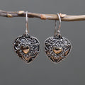 Heart-shaped earrings with intricate designs hanging on a branch against a gray background